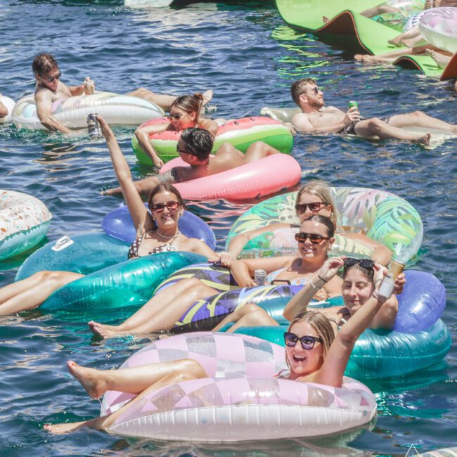 A group of people relaxing on colorful inflatable floaties in the water, smiling and waving, with boats in the background on a sunny day.