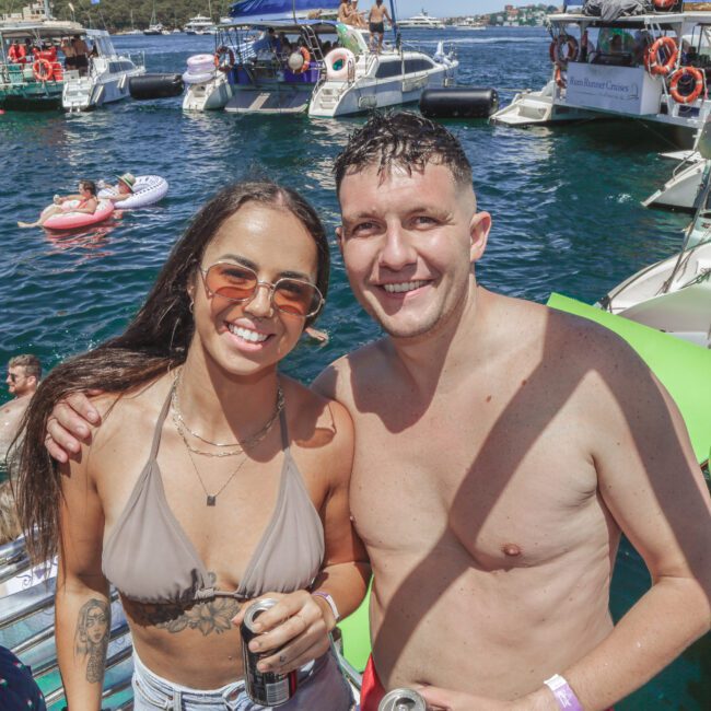 A smiling woman and man in swimsuits pose on a boat at a lively yacht party, with drinks in hand. Several boats and people swimming or lounging on floats are visible in the sunny, blue water background.