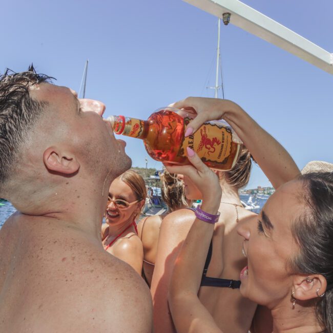 A woman pours Fireball whisky into a man's mouth at a lively outdoor boat party, with people swimming and socializing in the background under a clear blue sky.