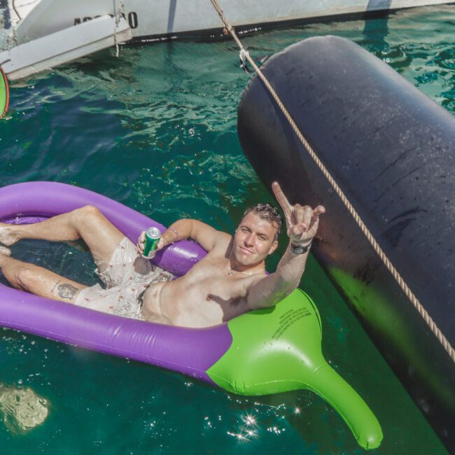 A man relaxes on a purple and green inflatable float in the water, holding a can and making a hand gesture, with others nearby and a boat in the background. The sun is shining and the water is clear and blue.