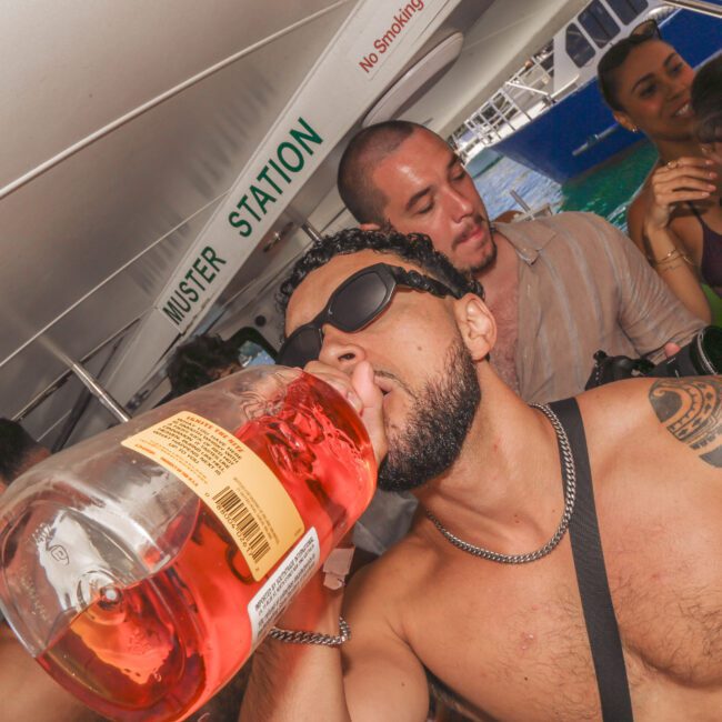 A shirtless man wearing sunglasses drinks from a large bottle of alcohol on a crowded boat. Several other people, some in swimwear, are gathered around, smiling and enjoying the lively atmosphere.