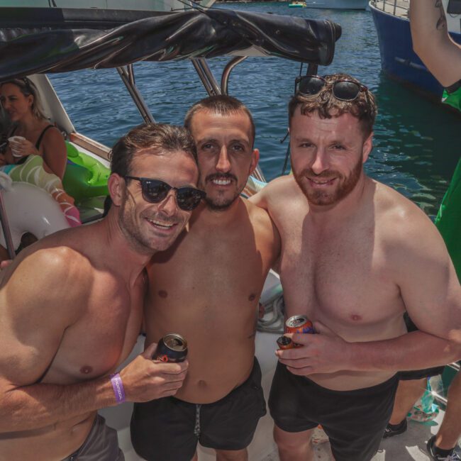 Three men in swim trunks smile and pose together on a boat, each holding a drink. Other people relax in the background, and boats float nearby on sunny blue water. The scene is lively and festive.