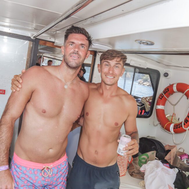 Two young men in swim trunks smile and pose together on a boat. One holds a drink can. There are bags, towels, and a "NO SMOKING" sign in the background, along with a life preserver on the wall.