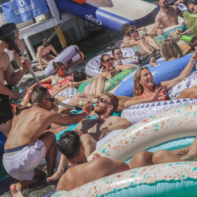 A group of people in swimsuits relax and socialize on inflatable pool floats in the water during a lively pool party, with one person dressed as a sailor entertaining the crowd.