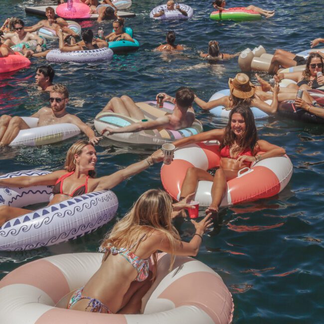 A group of people relax on colorful inflatable floats in a sunlit body of water, smiling and toasting drinks. The scene is lively and social, with many enjoying the summer weather. “Yacht Social Club” logo appears in the corner.