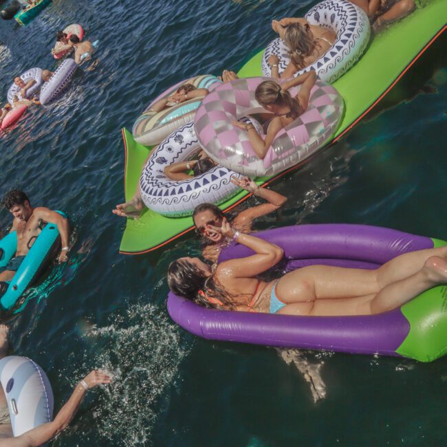 Several people relax on colorful inflatable pool floats in the water near boats on a sunny day. Most are wearing swimsuits, enjoying the social atmosphere. The scene is lively and festive.