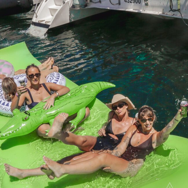 Three women in swimsuits relax on a green float in the water, smiling and posing for a selfie. One holds an inflatable crocodile while boats are docked in the background. Sunlight reflects off the water.
