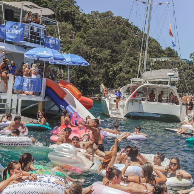 A large group of people enjoy a party on the water, floating on inflatable pool floats near boats. Some are climbing a slide on a large boat; lush green trees are visible in the background under a sunny sky.