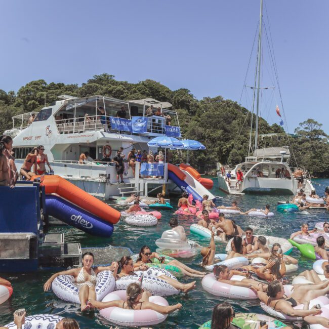A lively scene of people relaxing on inflatable pool floats in the water near two yachts, surrounded by trees and enjoying a sunny day. Many are in swimwear, talking, and having fun at a social event.