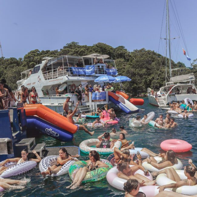 A large group of people relaxing on colorful inflatable floats in the water near docked yachts, with a blue sky and trees in the background at a lively summer party.
