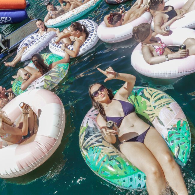 A group of people in swimsuits relax on colorful inflatable rings in a body of water, some holding drinks; one woman in the foreground smiles and makes a peace sign.