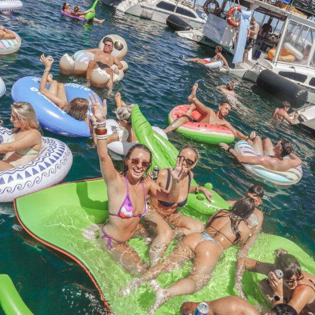 People relax on colorful inflatable floats in the water near several boats, enjoying a sunny day. Some are smiling, waving, or holding drinks, while groups of friends gather both in the water and on the boats.