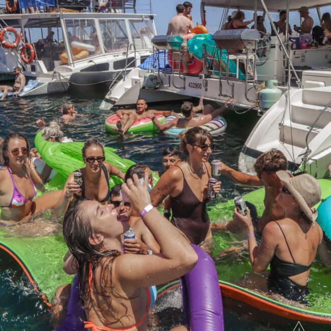 A group of people in swimsuits enjoy drinks and float on inflatable rafts in the water near anchored boats during a lively summer party. Some are cheering, laughing, and relaxing under the bright sun.