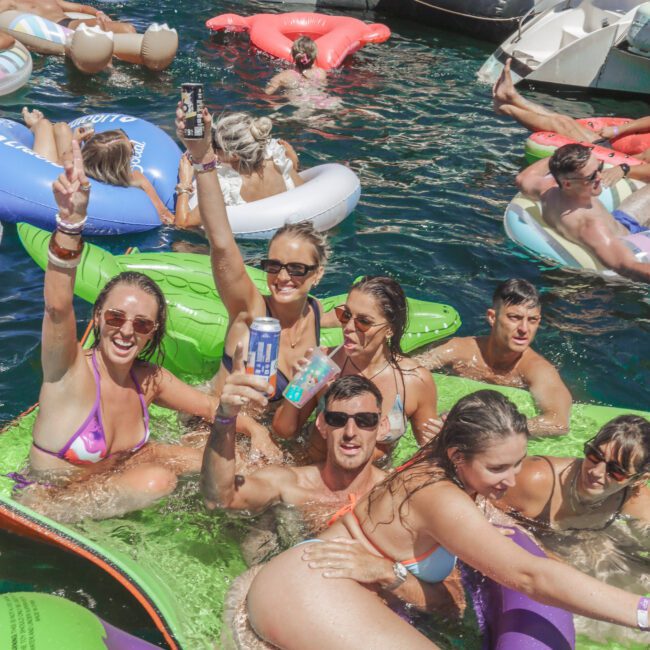 A group of young adults in swimsuits smile and pose with drinks while floating on colorful inflatables in the water near yachts during a lively party. The sun is shining and people are celebrating around them.