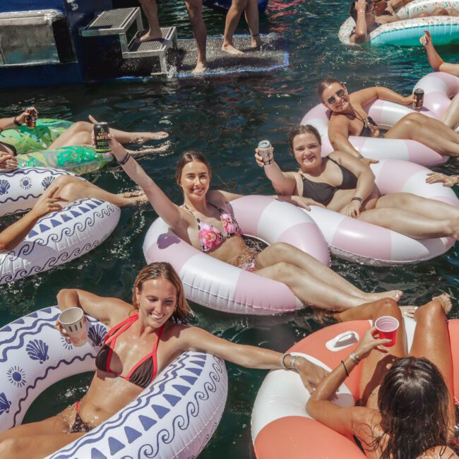 A group of women in swimsuits relax and smile on inflatable pool floats in the water, holding drinks and enjoying a sunny day together.
