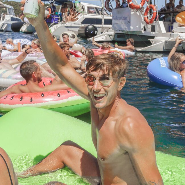 A smiling man with sunglasses holds up a drink while sitting on a green float in the water, surrounded by other people on floats and boats, enjoying a sunny day at a pool or boat party.