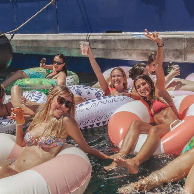Several women in swimsuits smile and pose while lounging on inflatable pool floats in the water at a lively outdoor party, with drinks in hand and others socializing in the background.