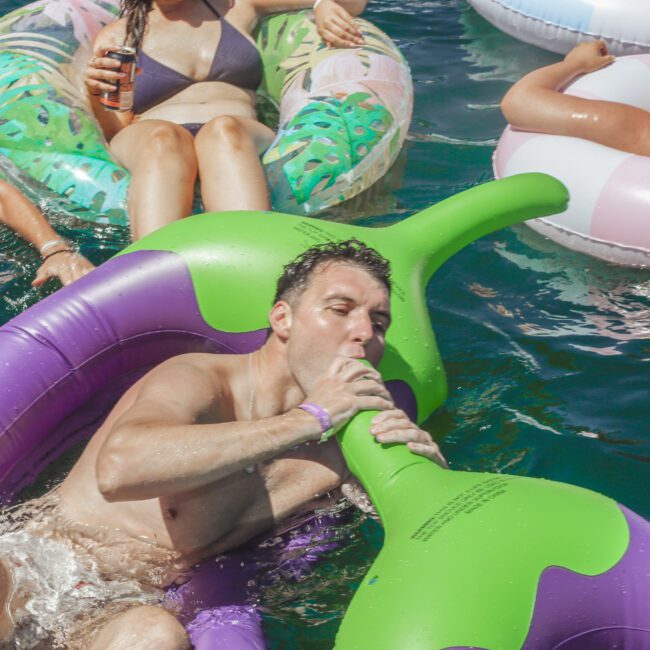 A man on a large, inflatable eggplant float appears to bite the stem while relaxing in a pool, surrounded by others on various inflatables. The scene is lively and sunny, suggesting a fun pool party.
