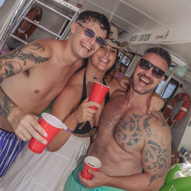 Three people in swimwear smile and pose with red cups on a boat, enjoying a sunny day. Two men with tattoos and a woman wear sunglasses. The boat’s interior and lifesavers are visible in the background.