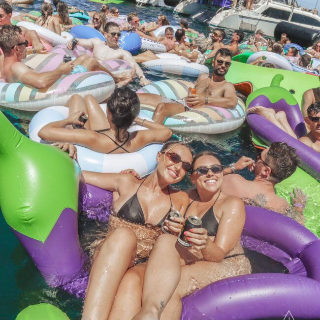A group of people in swimsuits relax on colorful inflatable floats in the water, laughing and holding drinks during a sunny yacht party. Boats are docked nearby, and the mood is lively and festive.