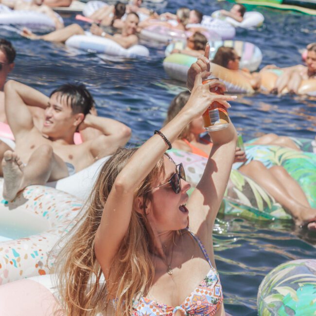 A woman in sunglasses and a floral bikini raises a drink while floating on an inflatable in a crowded pool or lake, surrounded by other people relaxing on floaties in the sun.