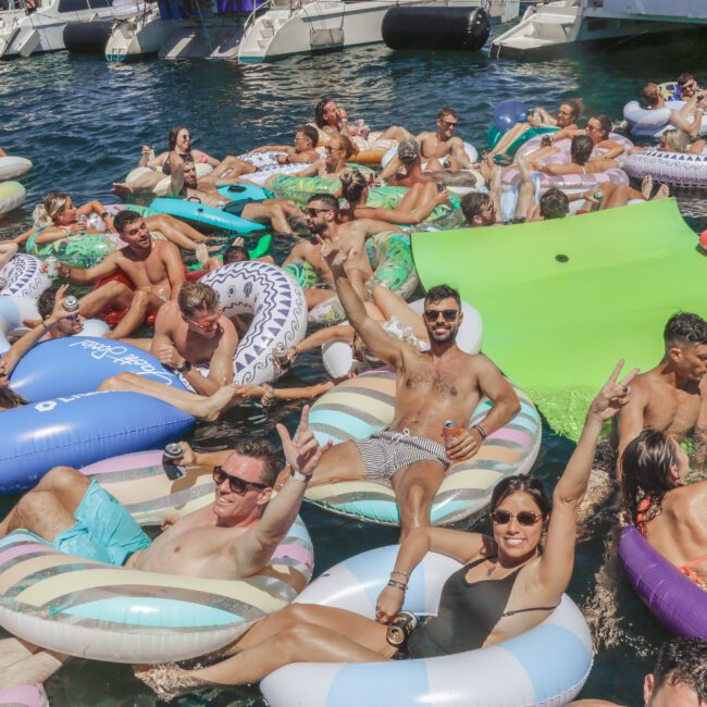 A large group of people relax on colorful pool floats in the water near several docked boats, enjoying the sunny weather and lively atmosphere at a boat party. Some people smile and make peace signs for the camera.