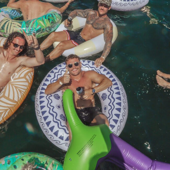 Three men with sunglasses and tattoos relax on colorful pool floats in a body of water, smiling and making hand gestures. Other people on floats are partially visible around them. The scene is sunny and lively.