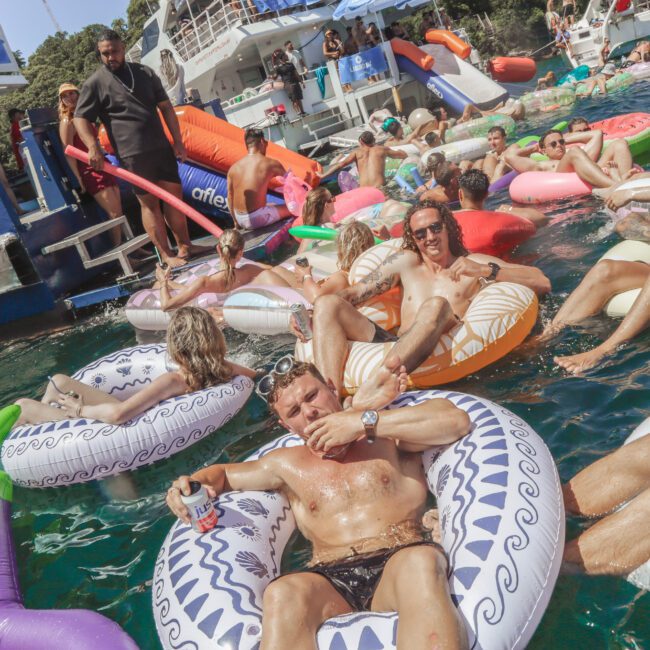 A group of people relaxing on colorful inflatable tubes in the water near docked boats on a sunny day, enjoying a lively outdoor gathering.