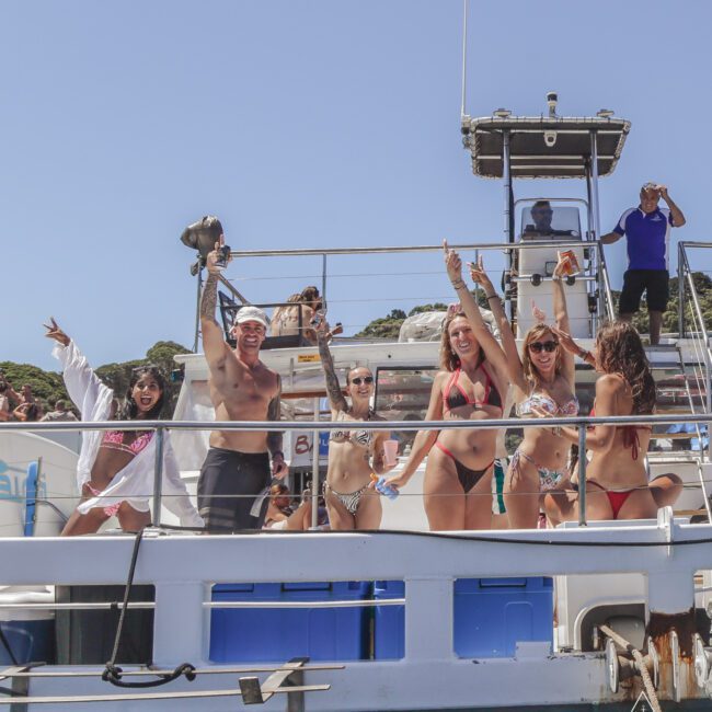 A group of smiling people in swimsuits pose and celebrate on the deck of a boat under clear blue skies, with greenery and a crew member in the background.