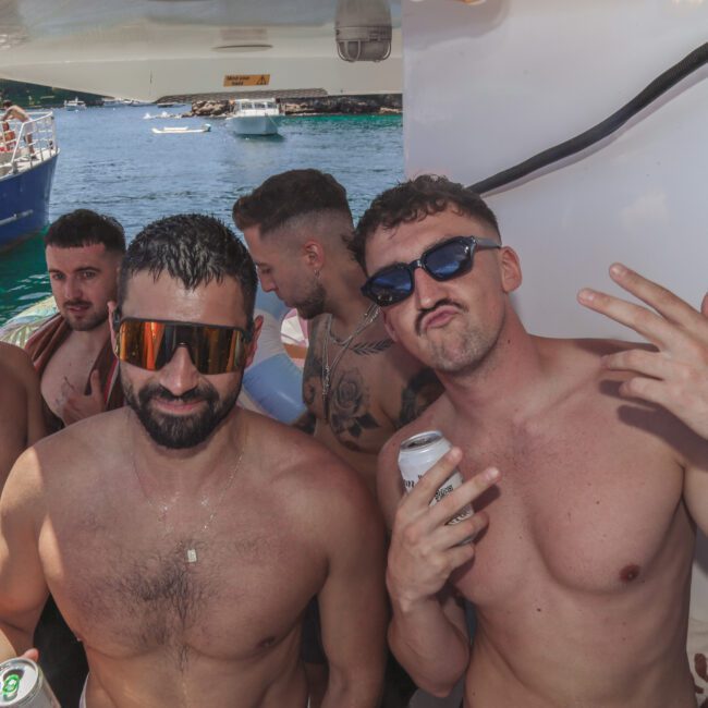 A group of shirtless men on a boat, holding drinks and posing for the camera. The background shows water, another boat, and a sunny day. One man makes a peace sign, while others smile and relax.
