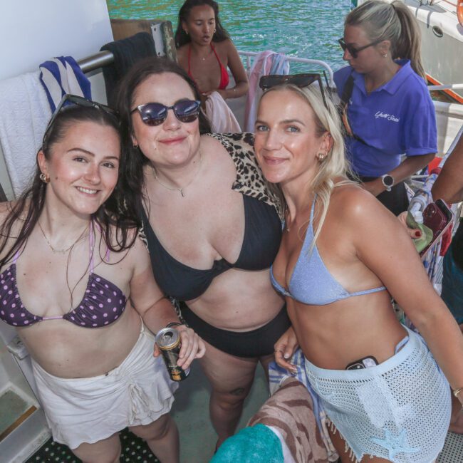 Three smiling women in swimsuits pose together on a boat, surrounded by others enjoying a sunny day by the water. The atmosphere is relaxed and festive, with towels and drinks visible.
