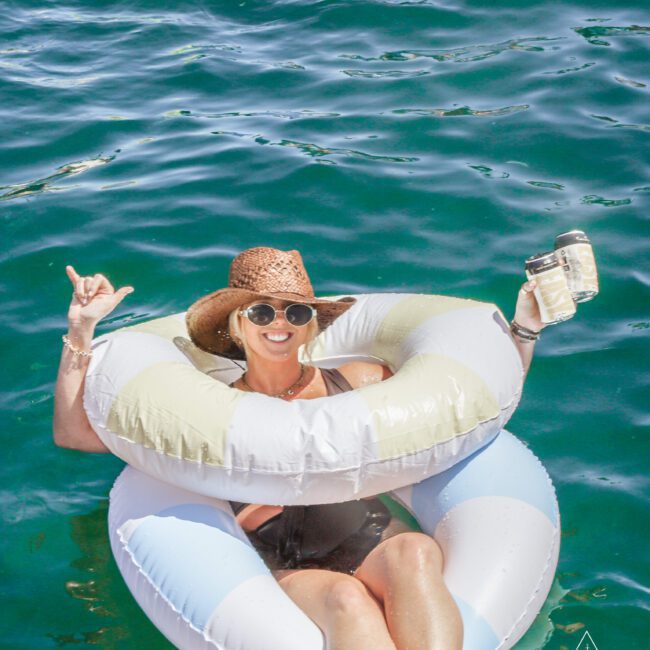 A woman in a sun hat and sunglasses floats on a blue-and-white pool tube in clear water, smiling and holding a drink jar, raising her hand in a "shaka" gesture.