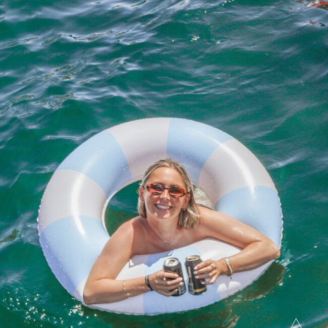 A smiling woman in sunglasses lounges in a blue and white inflatable ring on clear green water, holding two cans in her hands. The sunlight reflects off the water, creating a bright, cheerful scene.