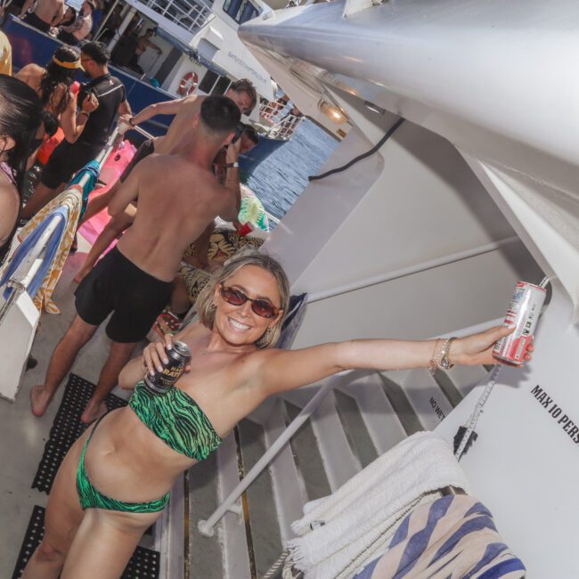 A woman in a green bikini smiles and raises a drink can on a crowded boat party, surrounded by people in swimwear, with the ocean and another boat visible in the background.