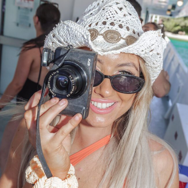 A smiling woman with long blonde hair, wearing a white crochet cowboy hat and sunglasses, holds a camera up to her face. She stands on a boat, with other people and water visible in the background.
