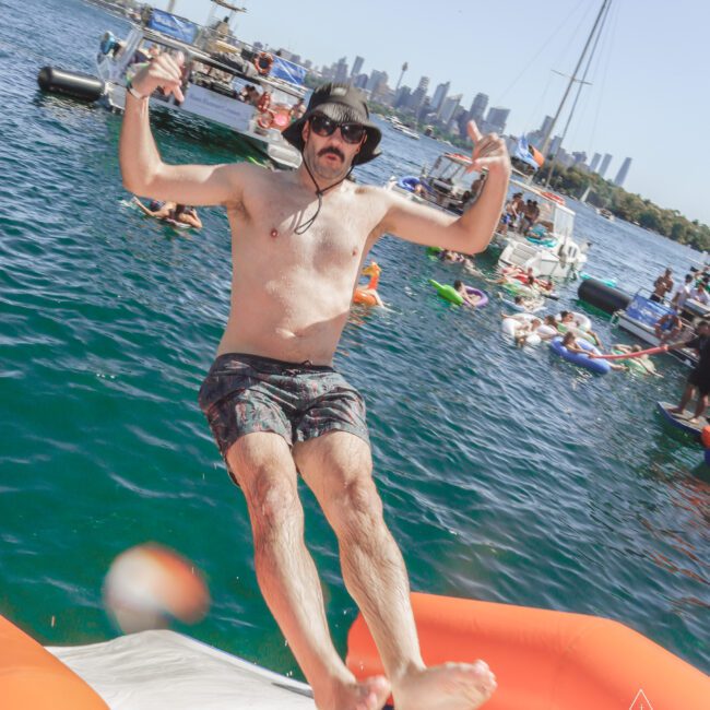 A man in swim trunks, sunglasses, and a hat falls backward off an inflatable float into the water, with boats and people enjoying a sunny day in the background.