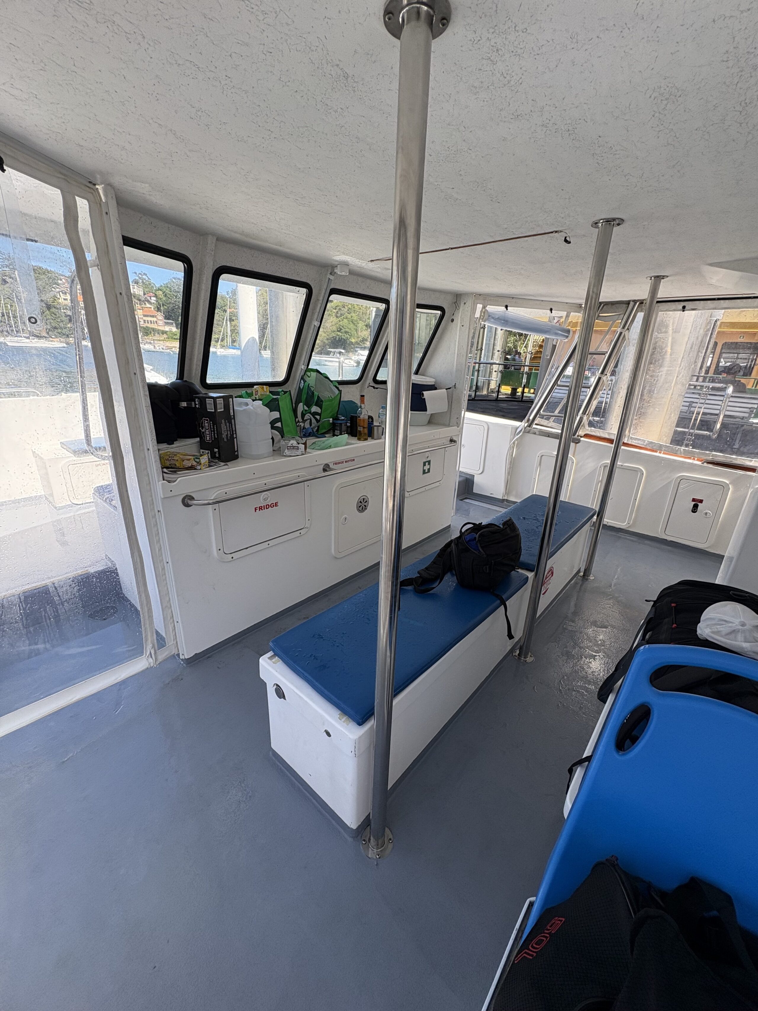 The interior of the Arcadia features white benches with blue cushions, metal poles, gear bags, and windows overlooking a marina. Sunlight brightens the clean, organized space inside this small boat.