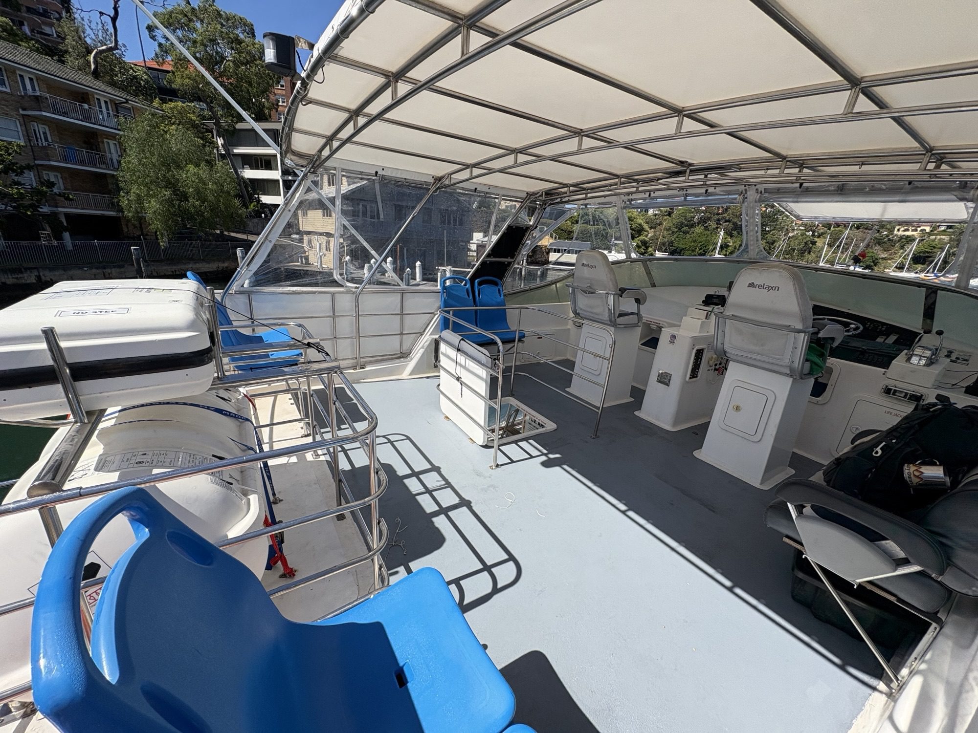 The Arcadia features an upper deck with blue and white seating, covered roof, railing, and control area with navigation equipment. Onshore buildings and trees are visible in the background.