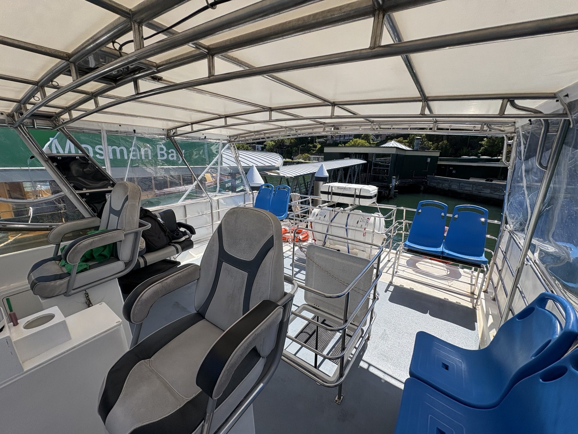 The upper deck of the Arcadia features empty seats, a steering area, and life rings, docked at a marina with water, signs, and other boats visible in the sunny background.