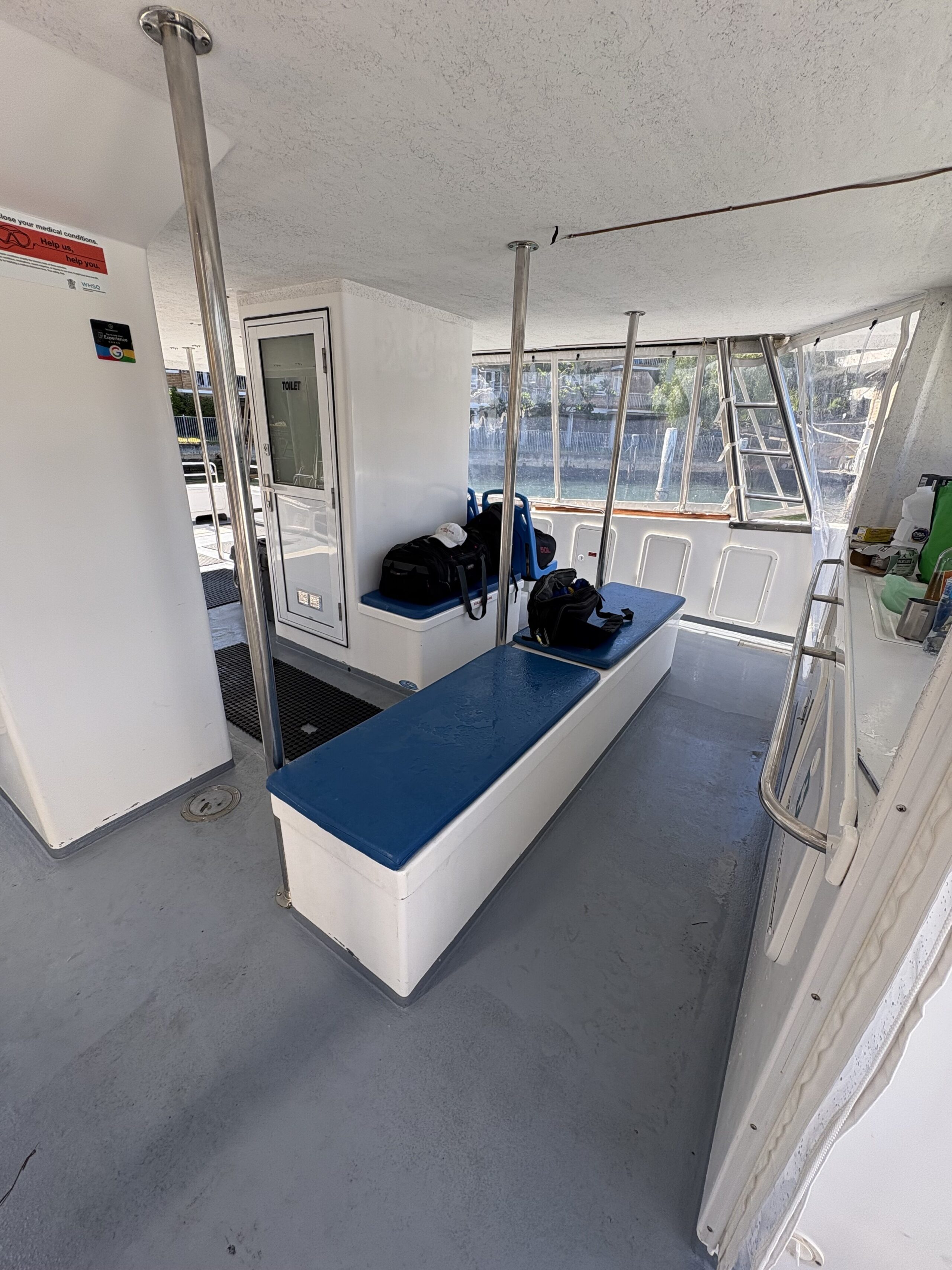 Interior of Arcadia boat featuring a blue-cushioned center bench topped with scuba diving gear, metal poles, white walls, and windows revealing water and a stone wall outside.