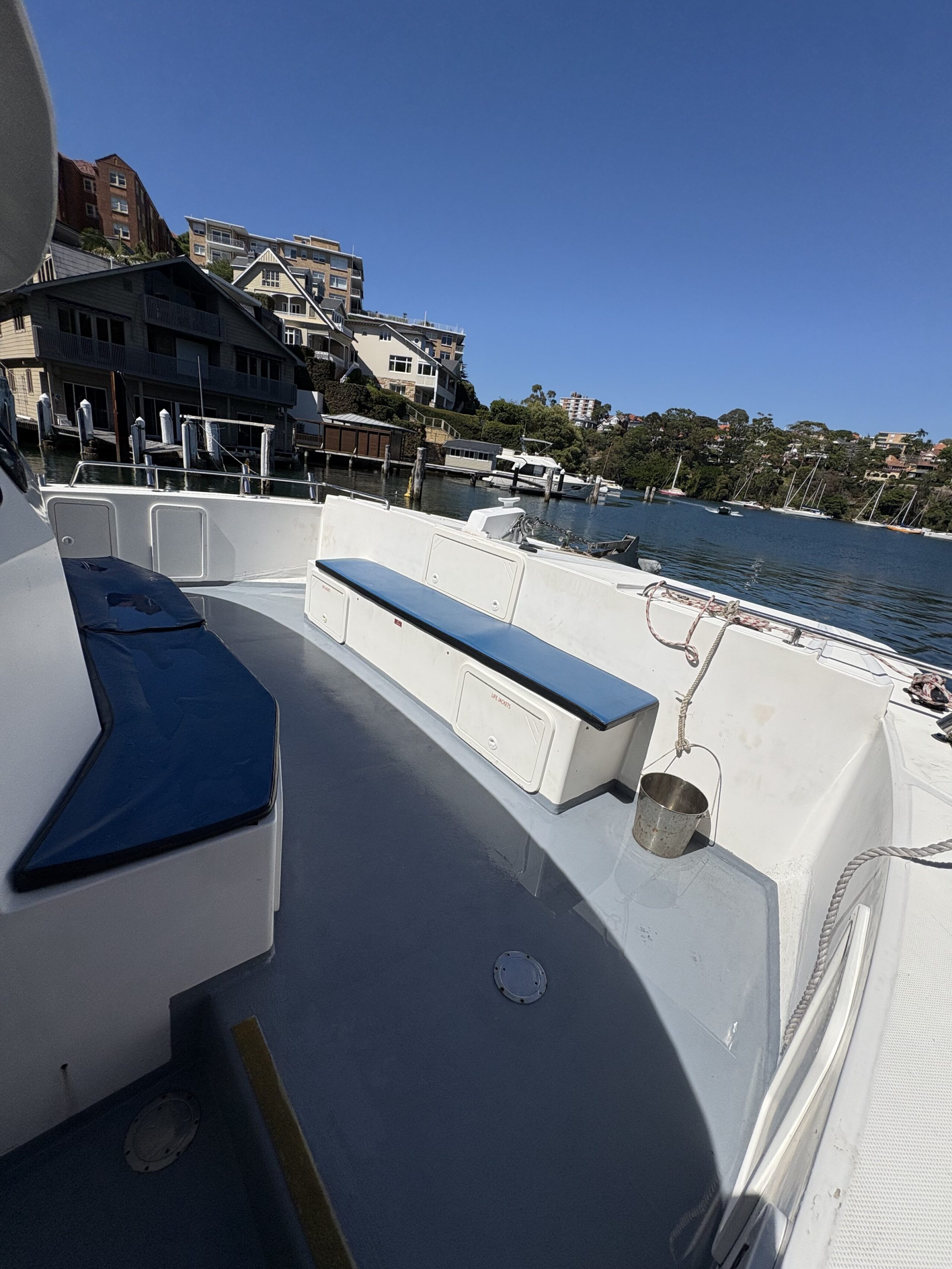 Enjoy the view from Arcadia’s deck, featuring blue bench seating with storage, docked near waterfront homes and trees under a clear blue sky and overlooking calm water.