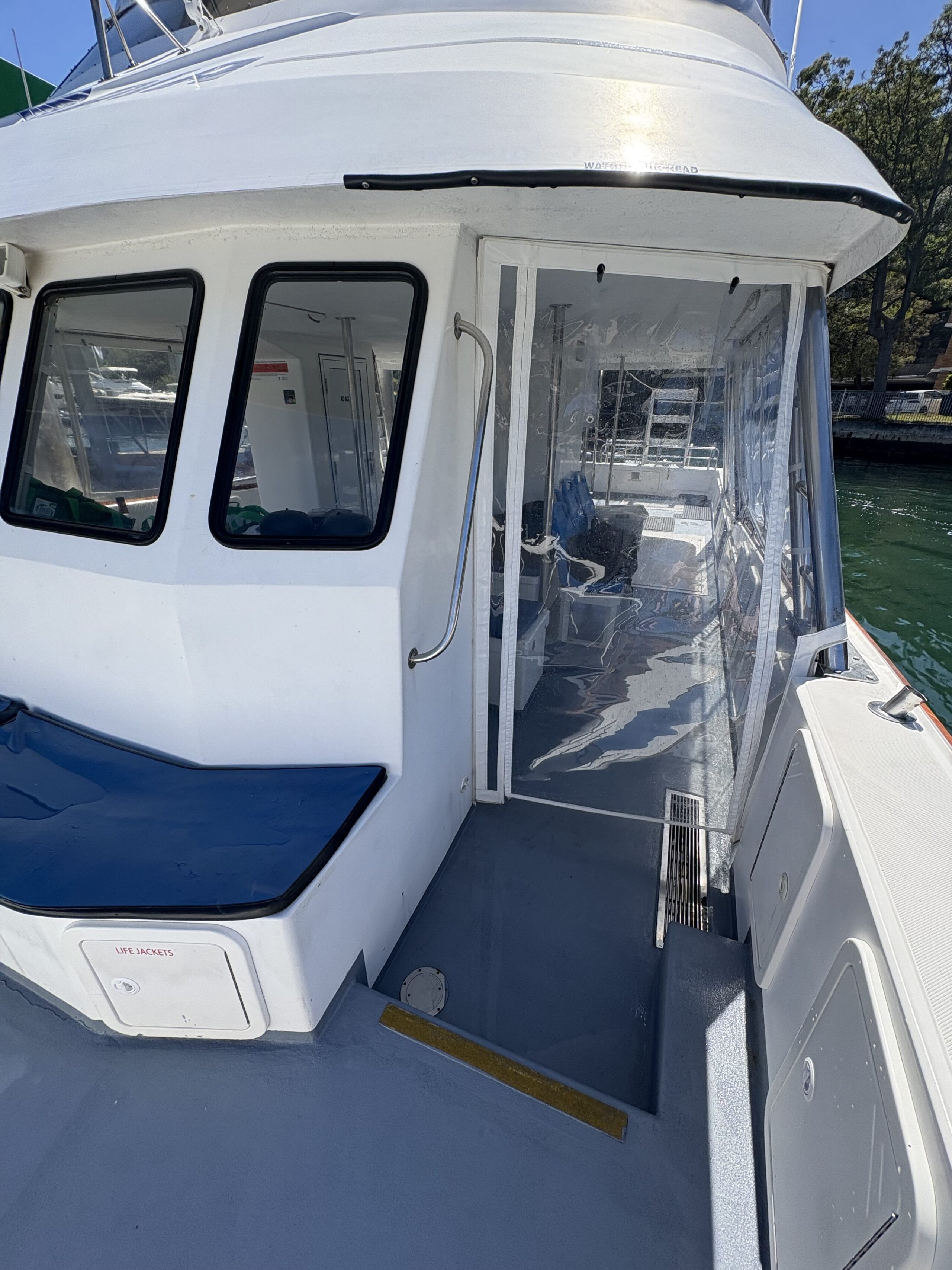 A view from Arcadia shows its blue bench on deck, a cabin door, clear plastic enclosure, and water-reflecting windows. Trees and water appear in the background.