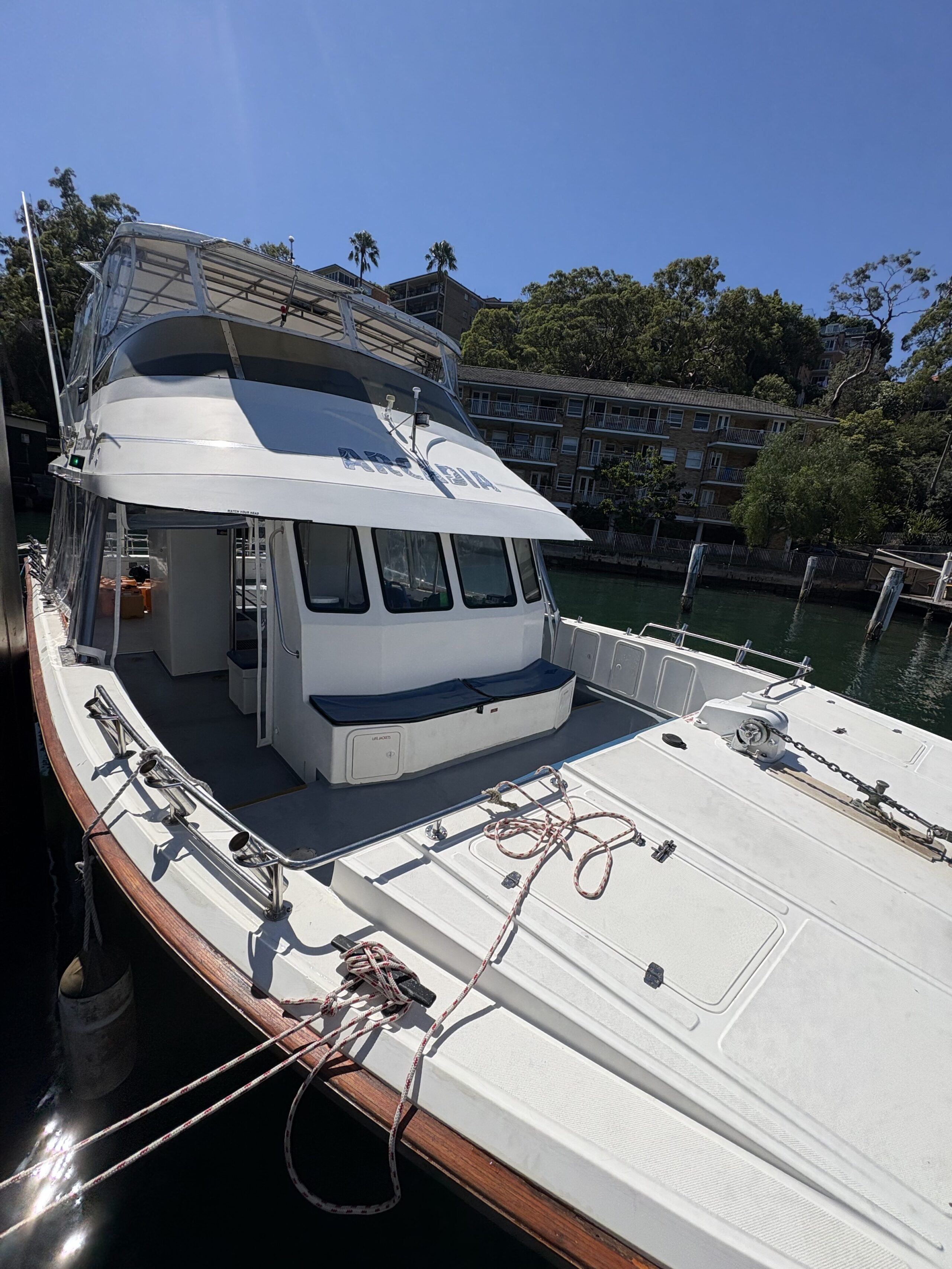 The Arcadia, a white motor yacht, is docked at a marina beside calm water. Ropes secure it to the pier, with trees and a multi-story building visible in the background on a sunny day.