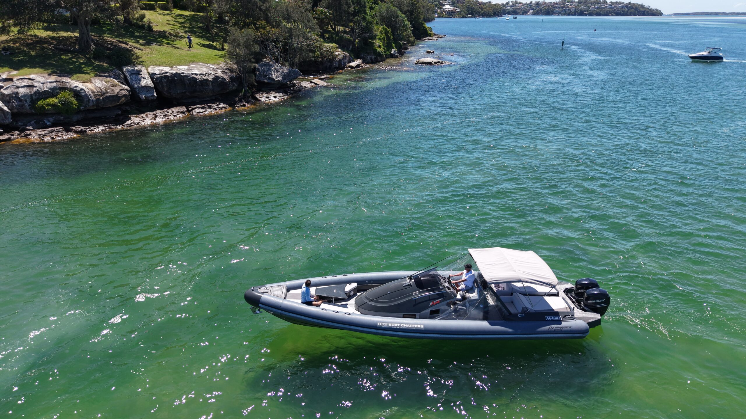 A small inflatable boat with a canopy floats on clear green water near a rocky shoreline with trees and grass. Another boat is visible in the distance under a sunny sky.
