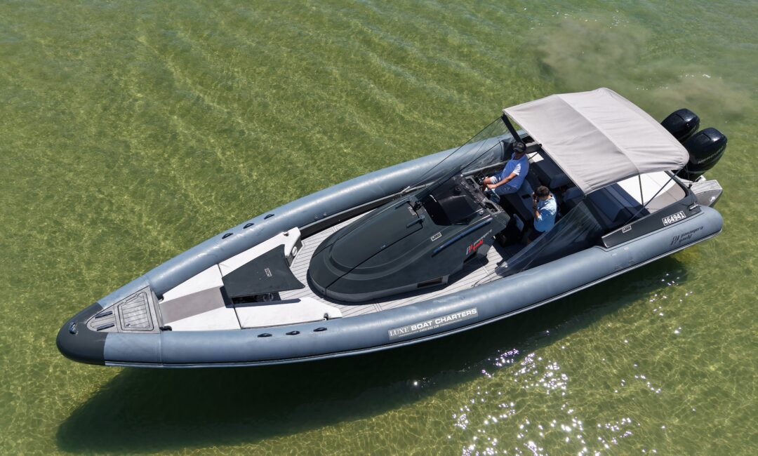 Aerial view of a gray inflatable motorboat with a canopy, two people seated at the helm, floating on clear shallow water. The boat has dual outboard engines and a sleek, modern design.