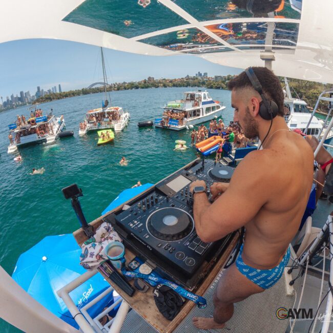 A DJ wearing swim trunks and headphones mixes music on a boat deck, overlooking a lively party on the water with people swimming, lounging on boats, and the city skyline in the background.