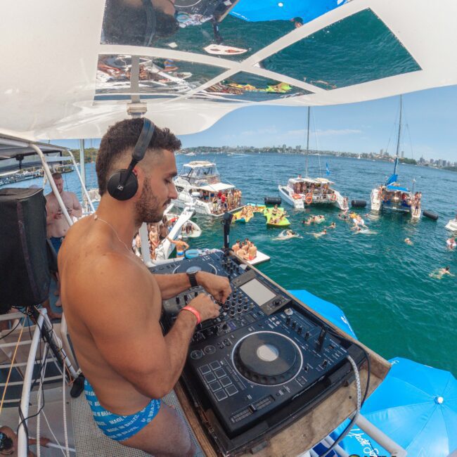 A man in blue swim trunks DJs on a boat, wearing headphones. Several boats with people partying and swimming are anchored nearby on a sunny day, with blue umbrellas and calm water in the background.