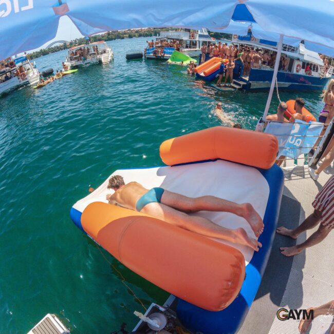 A man in swim briefs lies face down on an inflatable float by the edge of a boat, surrounded by people partying on nearby boats and platforms in a sunny, lively scene on the water.