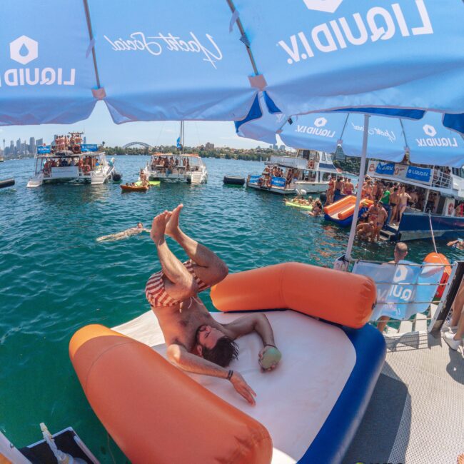 A man lounges playfully on an inflatable mattress under umbrellas on a boat, surrounded by people enjoying a lively party on the water with other boats and swimmers nearby under a sunny sky.