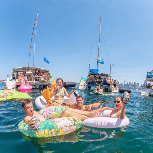 A group of people relax on colorful inflatables, drinking and smiling, in the water near anchored boats on a sunny day. The city skyline is visible in the background under a clear blue sky.